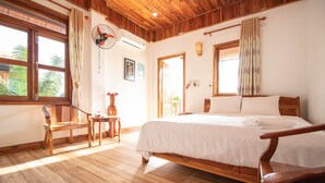 Un dormitorio con piso de madera, una cama, una silla, una mesita y un ventanal con cortinas.
