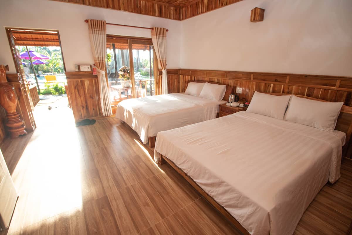 A hotel room with two beds, wooden flooring, a large window with curtains, and a view of the outdoors.