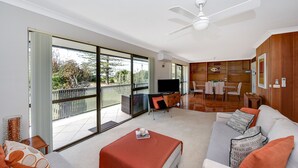 A modern living room with a sofa, a coffee table, and a TV. There is a balcony with a view of trees and a fence.
