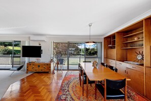 A spacious dining area with a wooden table and chairs, a large window with a view of a garden, and a television mounted on the wall. - Fi s Relaxing Beach House directly opposite dog beach (Port Macquarie)