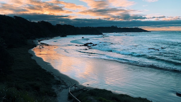 A coastal landscape with a curved shoreline, rocky outcrops, and a clear sky with clouds.