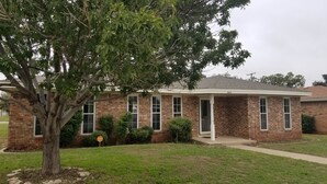 A single-story brick house with a covered porch, a tree in front, and a well-maintained lawn.