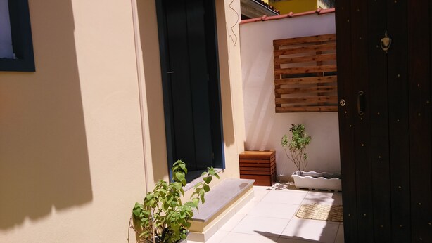 Un estrecho pasillo exterior con suelo de baldosas, un banco de madera, una mesita y una planta en maceta.