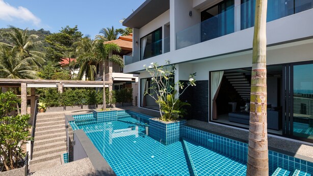 Een modern huis met twee verdiepingen, met een zwembad, omringd door tropische begroeiing en palmbomen.