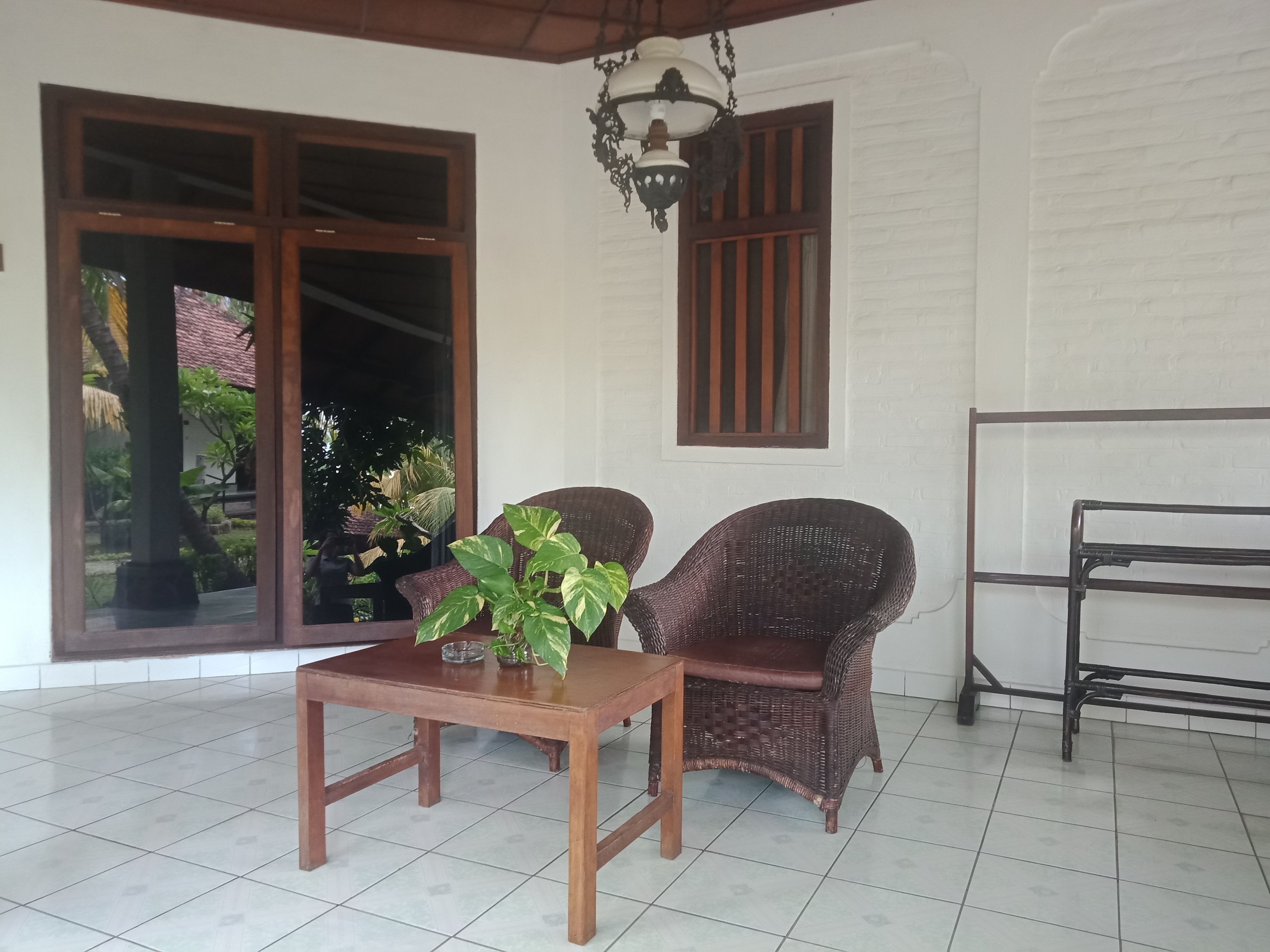 Una habitación con una mesa de madera, dos sillas de mimbre, una lámpara colgante y un ventanal con persianas verticales.