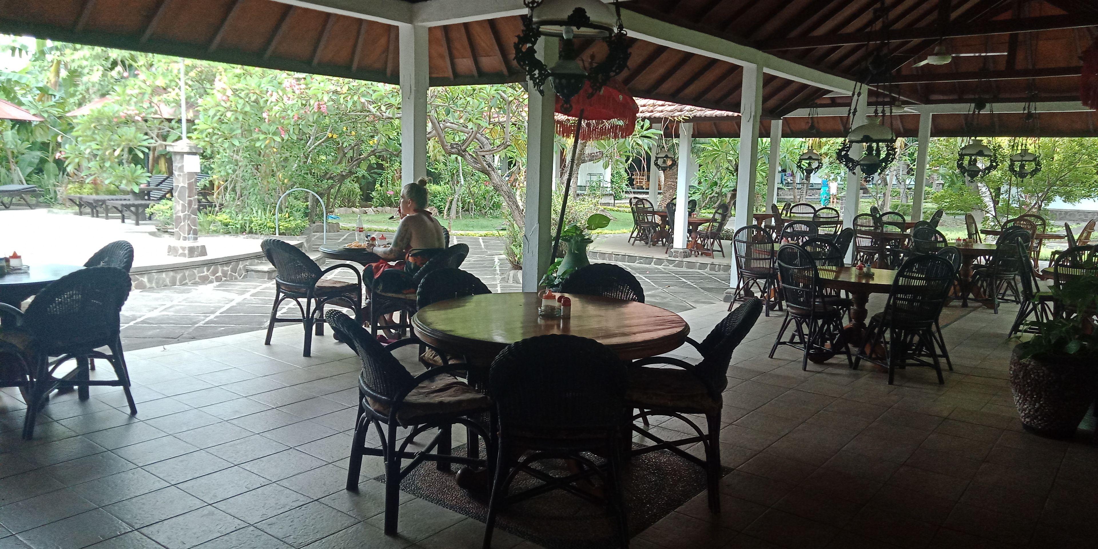 An outdoor covered dining area with multiple tables and chairs, surrounded by greenery.