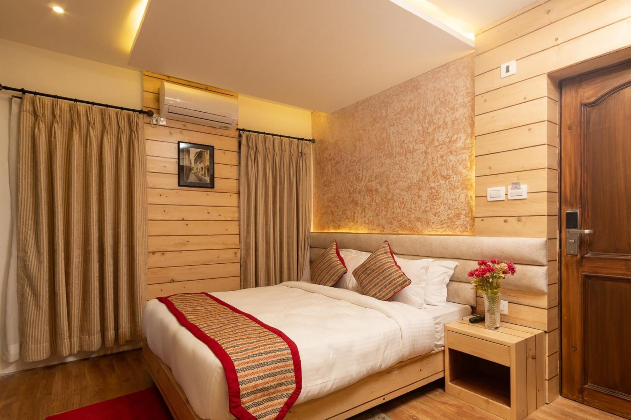 Un dormitorio con cabecera de madera, cama con ropa de cama blanca y almohadas a rayas, mesita de noche con un jarrón de flores y puerta de madera.