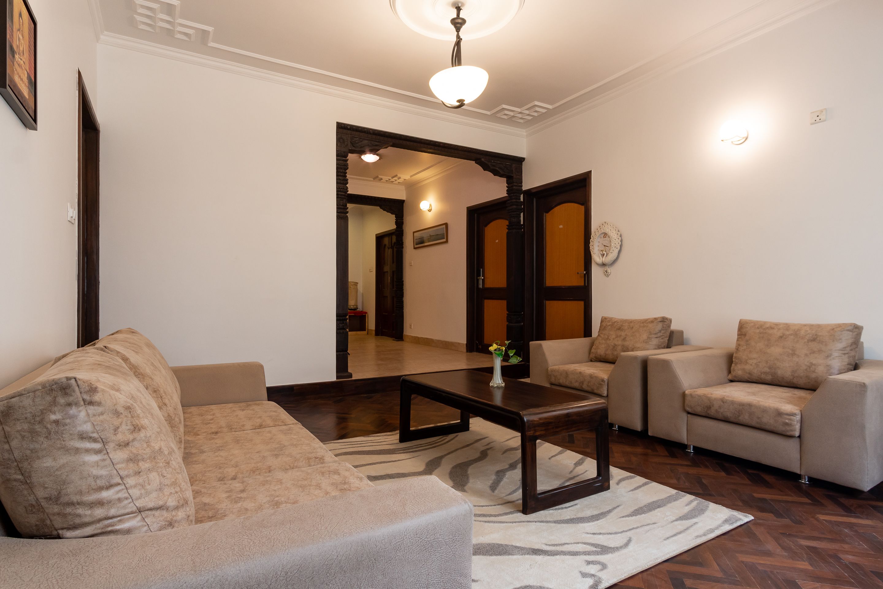Un salón con un sofá, sillones, una mesita de centro y un tapete estampado. Hay una puerta que conduce a otra habitación y una lámpara fijada a la pared.