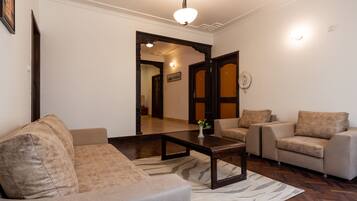 Un salón con un sofá, sillones, una mesita de centro y un tapete estampado. Hay una puerta que conduce a otra habitación y una lámpara fijada a la pared.