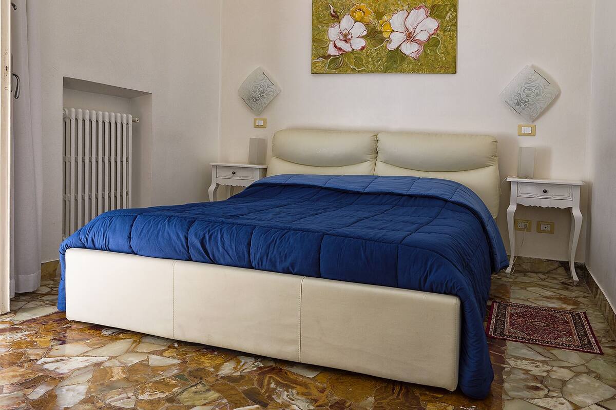 Quarto com cama, mesas de cabeceira, radiador e pintura floral na parede.