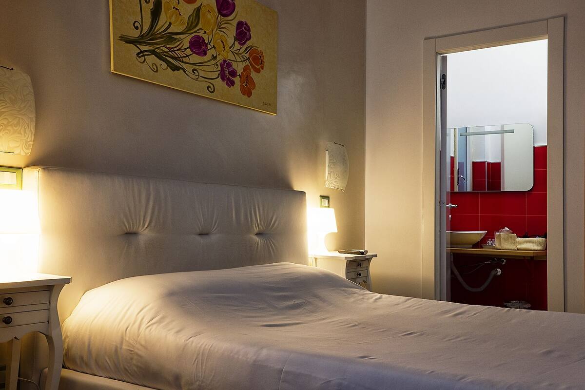 Quarto com uma cama grande, mesa de cabeceira com abajur, quadro na parede e porta para um banheiro com parede de azulejos vermelhos e espelho.
