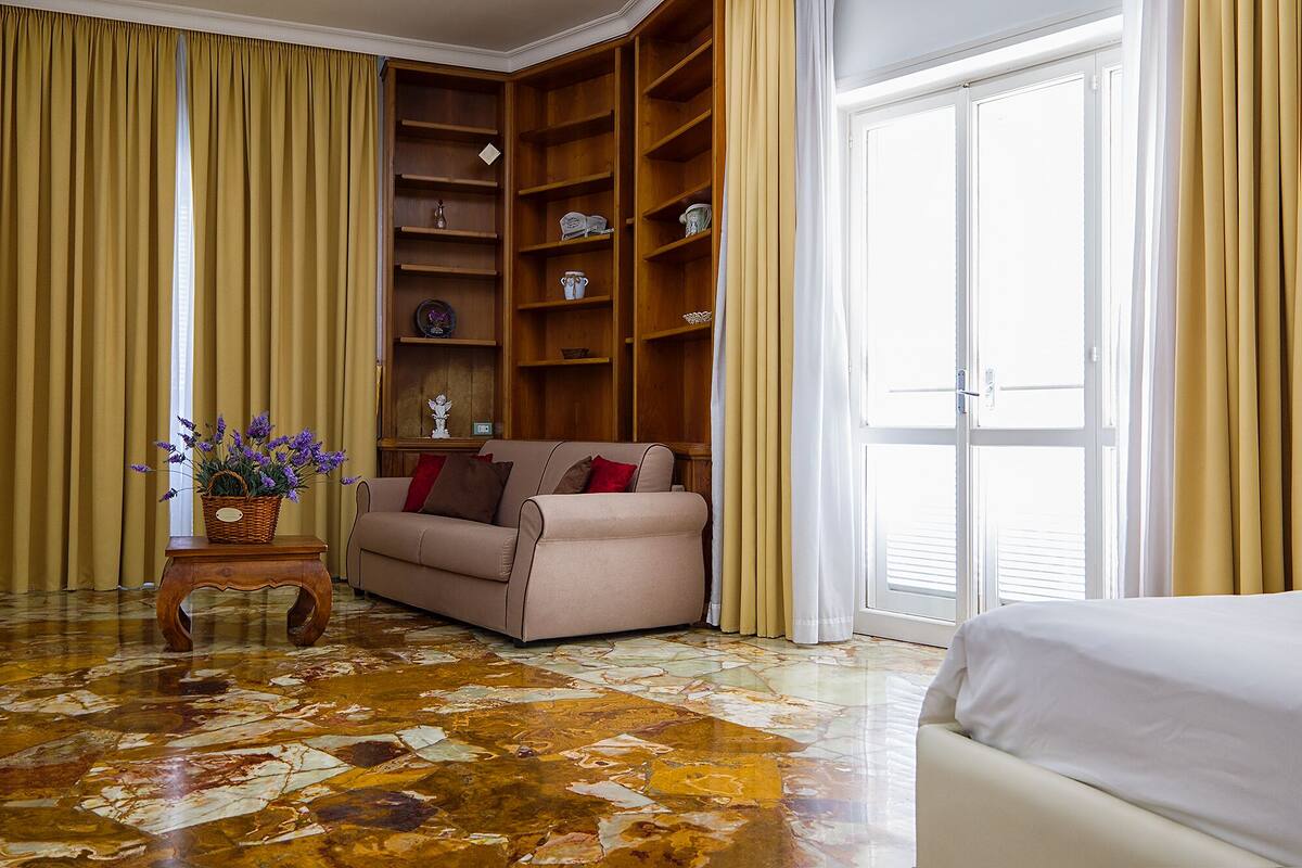 Um quarto com sofá bege, estante de madeira, piso de mármore e uma grande janela com cortinas.