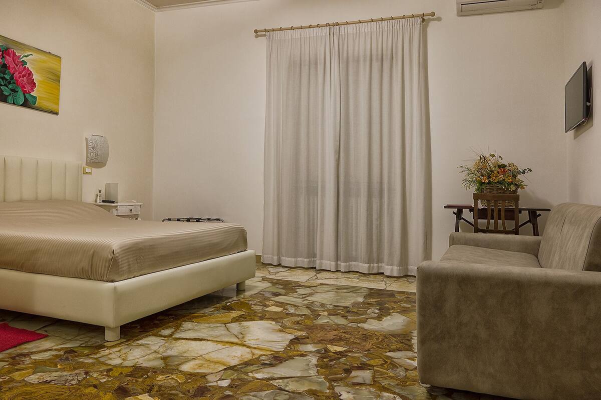 Quarto de hotel com cama, uma cadeira, uma mesinha de centro com um vaso e um quadro na parede.