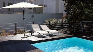 Um deck de madeira com duas espreguiçadeiras e uma mesa, uma piscina pequena e um guarda-sol branco.