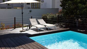 Eine Holzterrasse mit zwei Liegestühlen und einem Tisch, ein kleiner Pool und ein weißer Sonnenschirm.