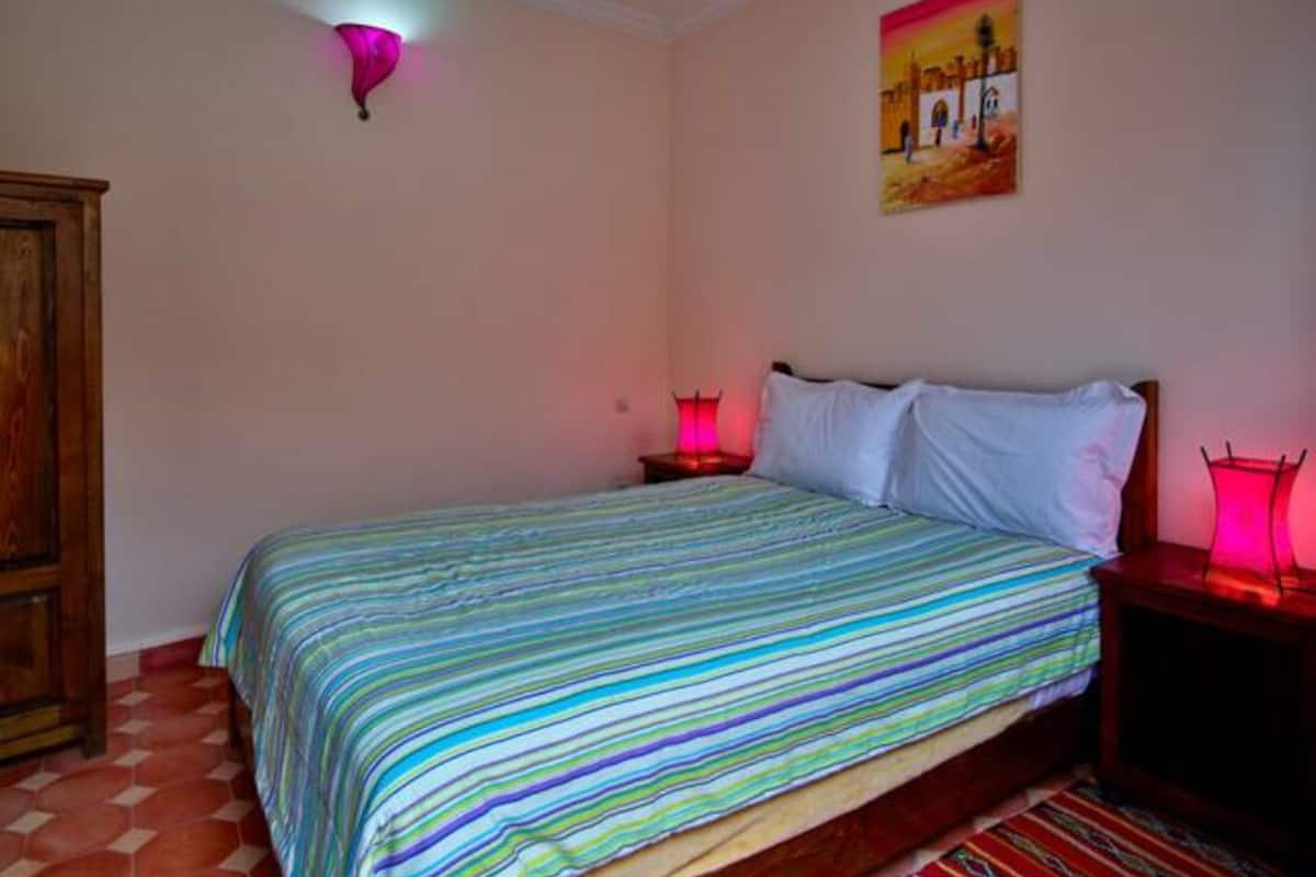 A bedroom with a bed, a nightstand, a lamp, a painting on the wall, and a wooden wardrobe.