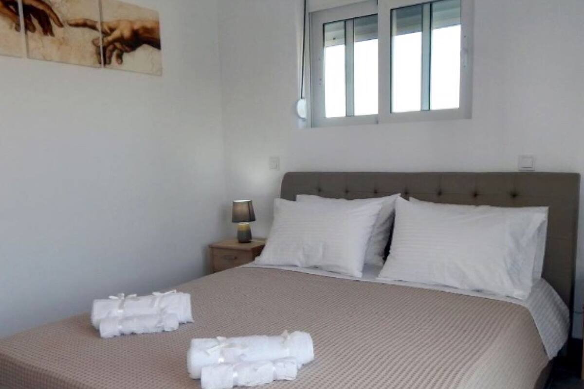 A neatly made bed with a beige bedspread and pillows, a nightstand with a lamp, and a window with blinds.