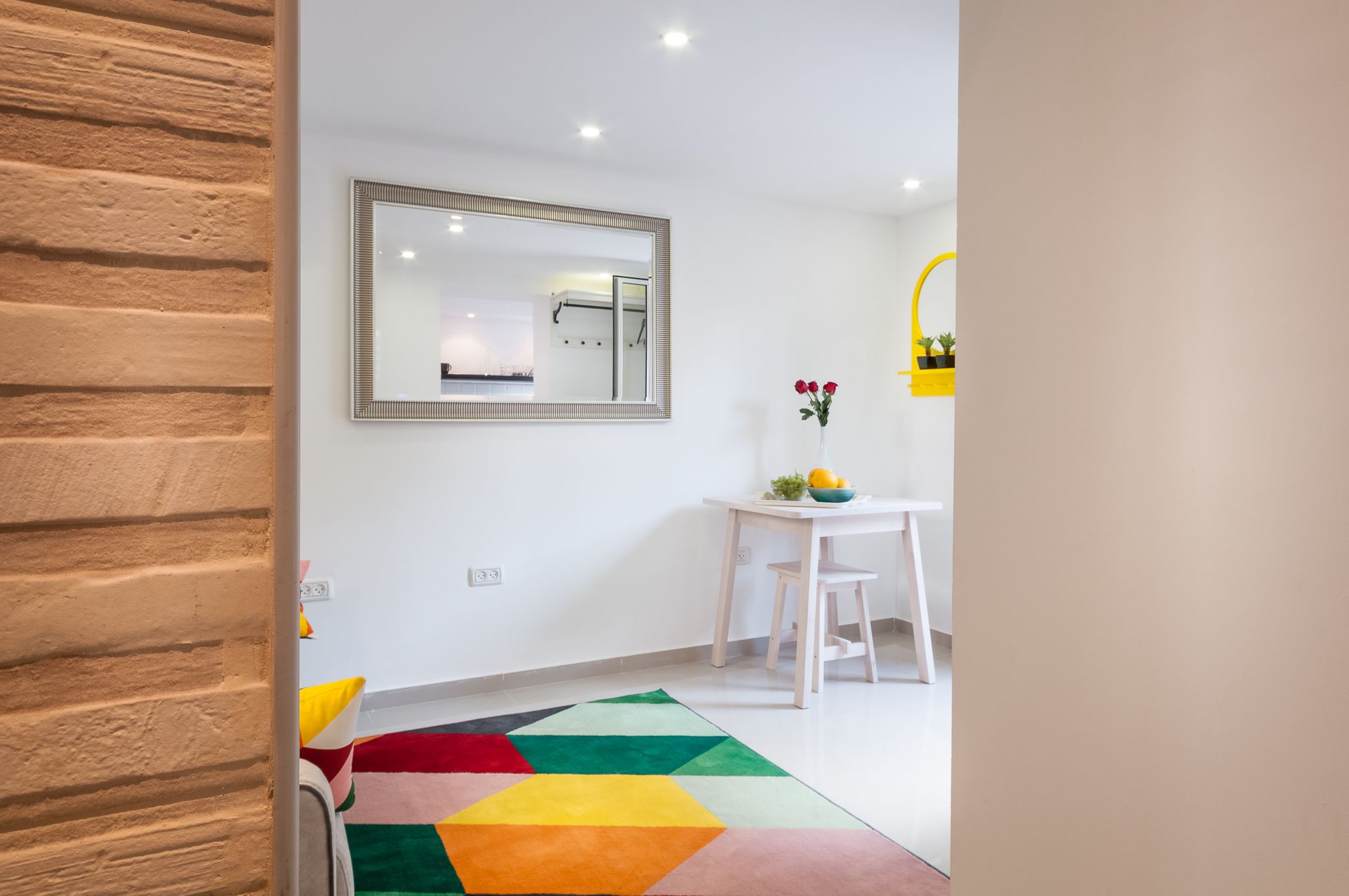 Een moderne, goed verlichte kamer met een witte wand, een grote spiegel, een wit bureau met stoel en een kleurrijk vloerkleed.