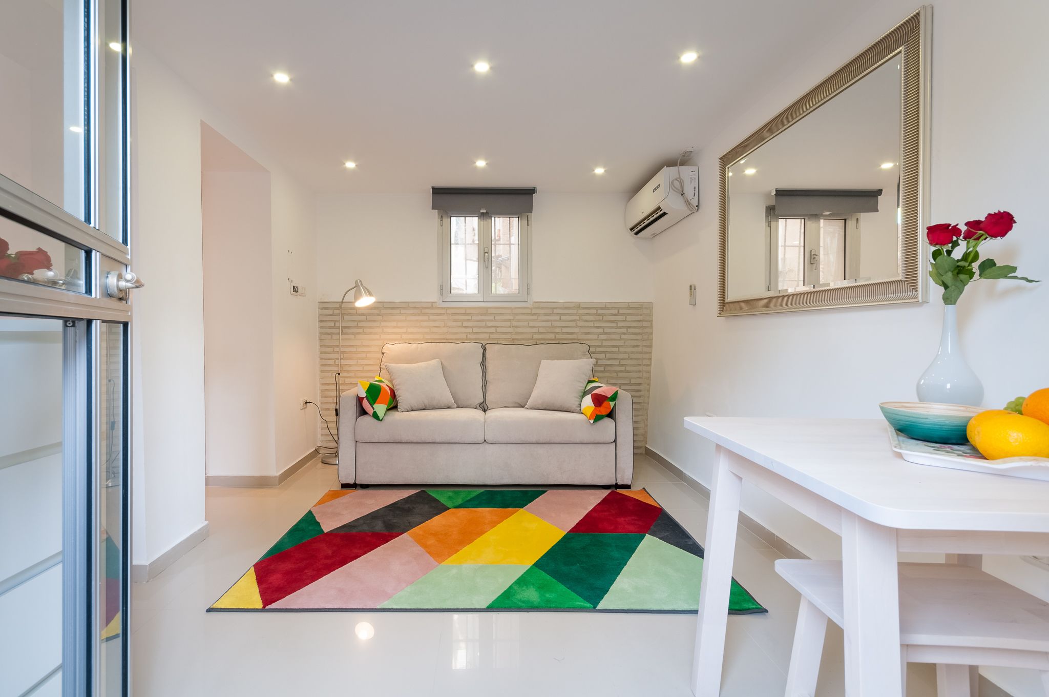 Un soggiorno moderno con un divano, un tappeto colorato, un tavolo bianco e un vaso con fiori.
