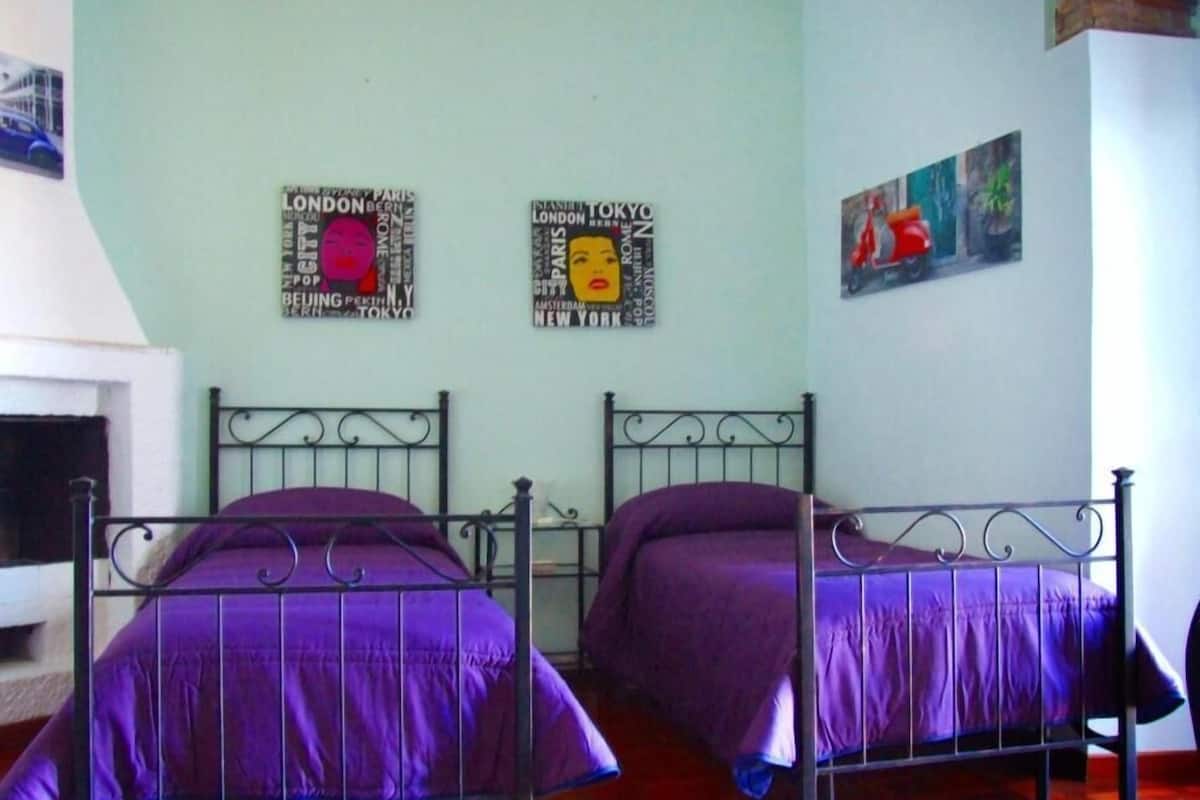 A room with two metal beds with purple bedding, a small table, and a wall with travel-themed decorations.