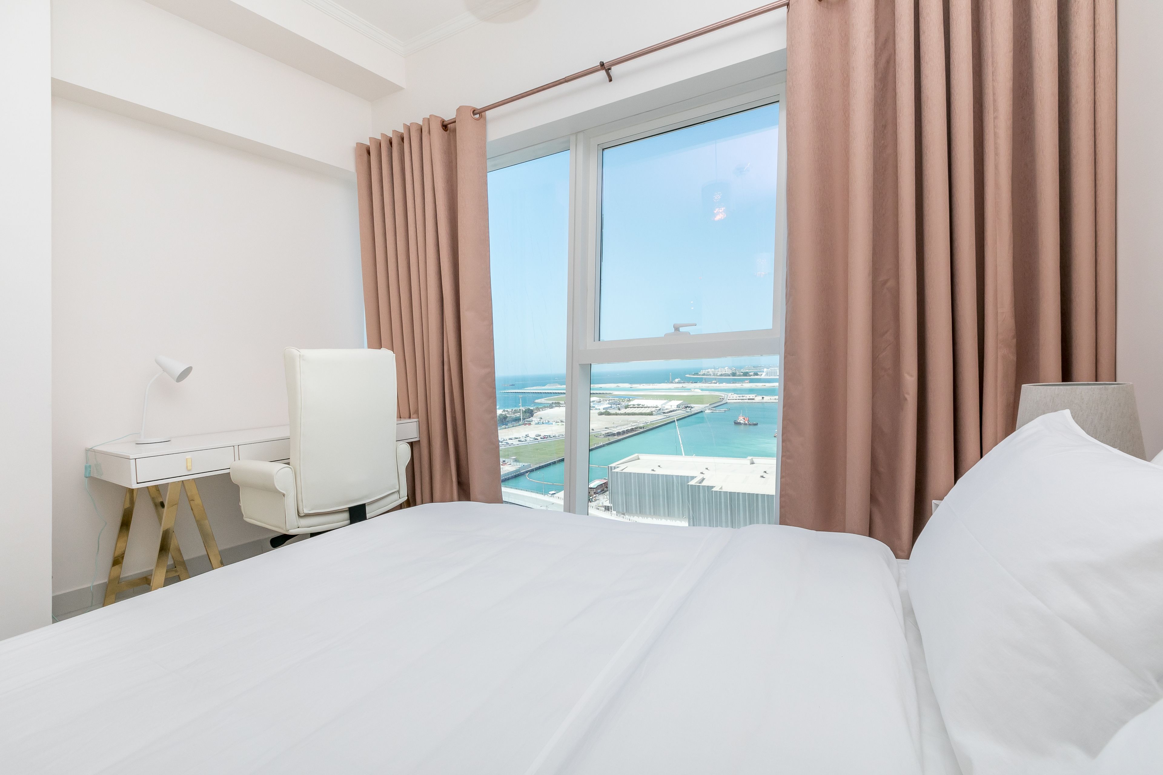A hotel room with a bed, a desk, a chair, and a large window offering a view of a port and buildings.