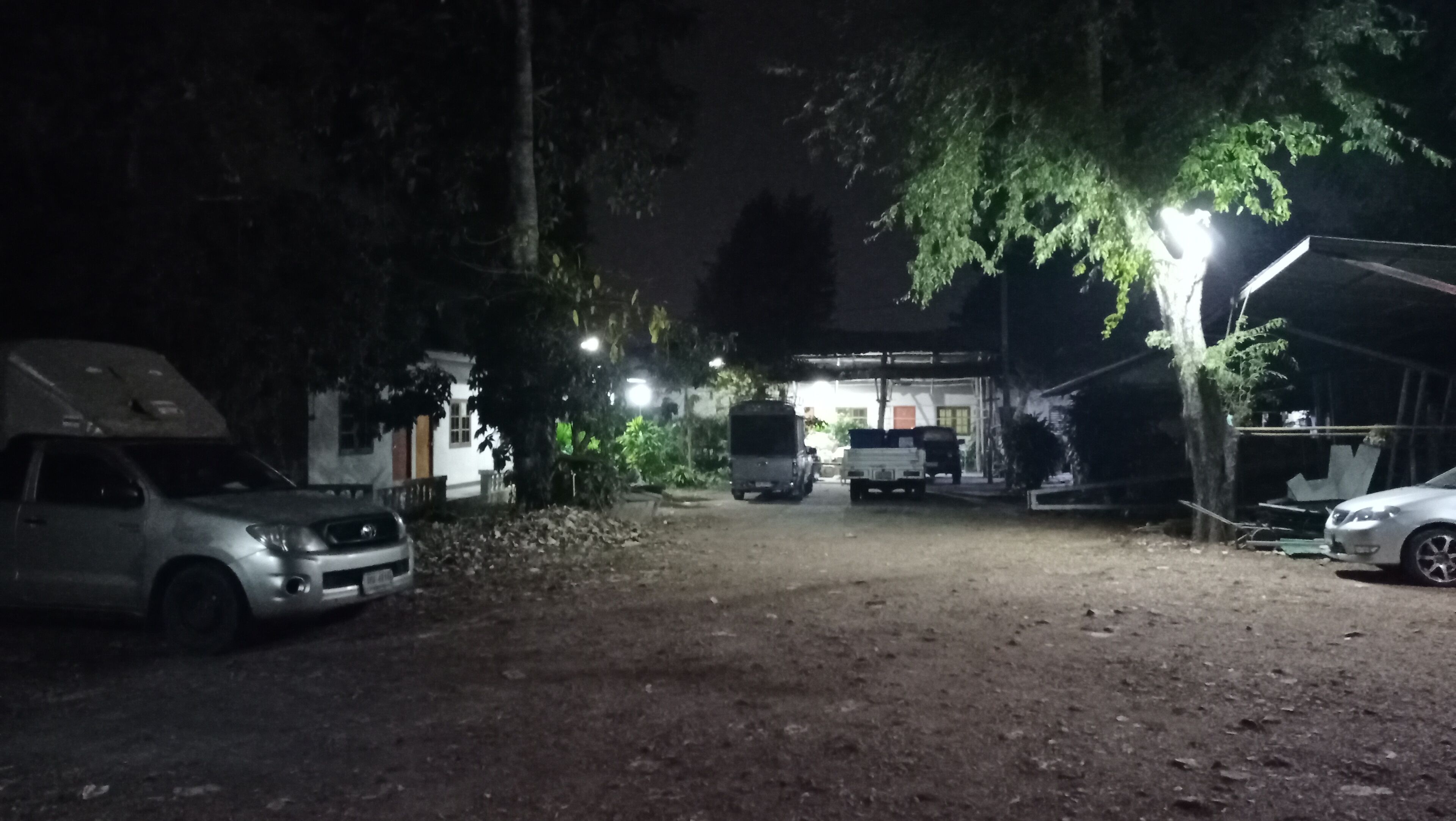 A nighttime scene with a dirt ground, parked cars, and a building with a lit window.