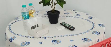 Una mesa redonda con mantel floral, una planta en maceta, un control remoto y una silla de plástico.