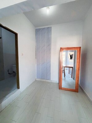 A room with a tiled floor, a vertical striped curtain, a wooden-framed mirror, and a visible bathroom with a shower. - Cozy Residence Wedasari Bali (Denpasar)