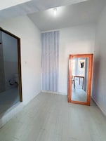 Una habitación con piso de baldosas, cortina a rayas verticales, espejo con marco de madera y un baño con ducha a la vista.