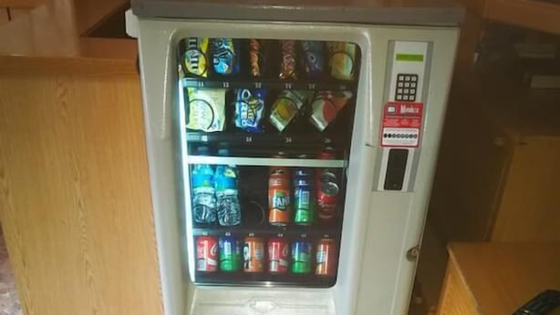 Un distributeur automatique proposant diverses boissons, situé dans une pièce dont le sol est carrelé et qui est équipée de meubles en bois.