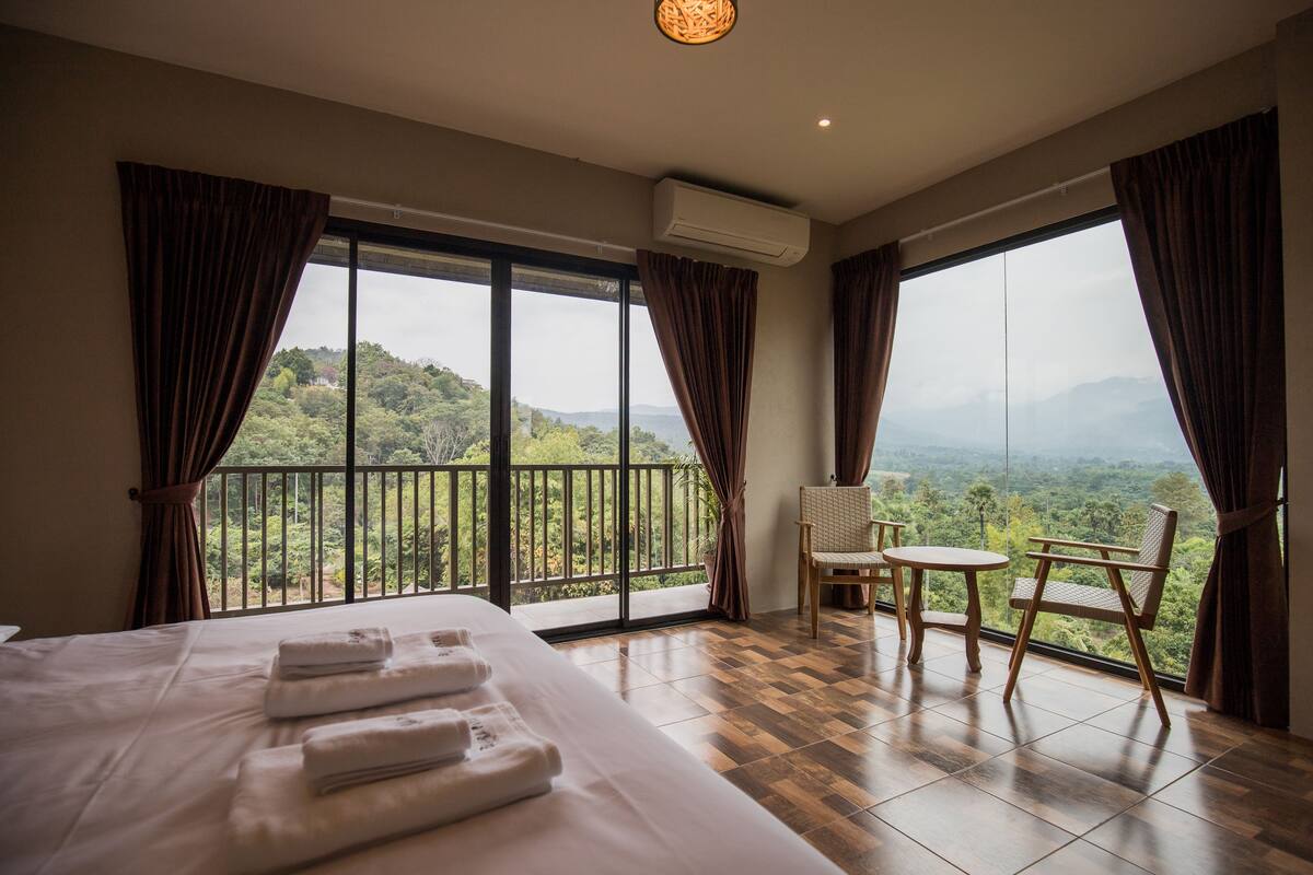 A hotel room with a large bed, a small table, two chairs, and a balcony with a view of the mountains.