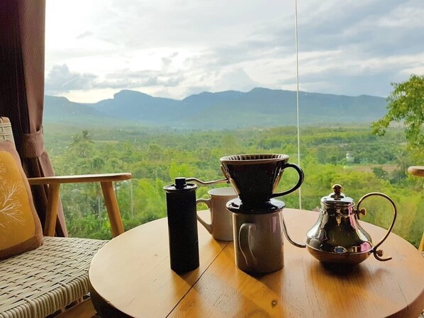 Una mesa redonda de madera con una cafetera, una taza y un termo en un balcón con vistas a un paisaje montañoso.