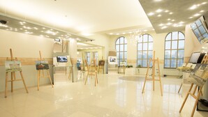 Une galerie d’art spacieuse, dotée de plusieurs chevalets sur lesquels sont exposées des peintures, de grandes fenêtres laissant entrer la lumière naturelle et d’un aménagement intérieur moderne et épuré.