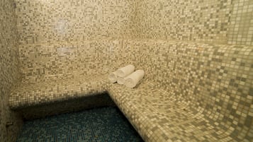 A corner of a room with mosaic tile walls and floor, featuring a curved bench with rolled towels on it.