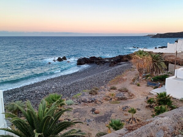 A coastal view with a pebbly beach, rocky shoreline, and a white building with a balcony overlooking the sea.
