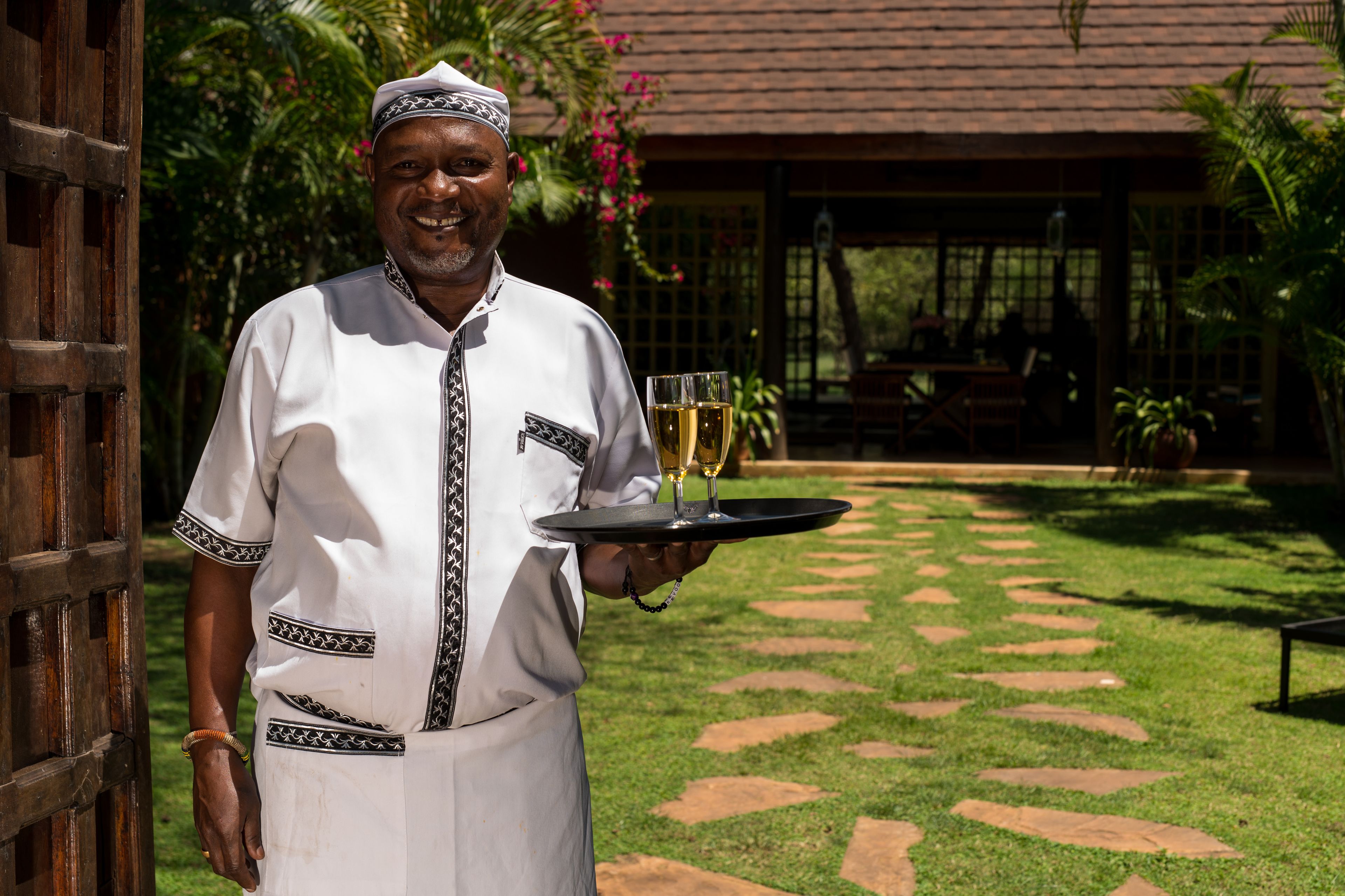 Uma pessoa de uniforme branco segura uma bandeja com uma taça de vinho, em um jardim com um caminho e plantas tropicais.
