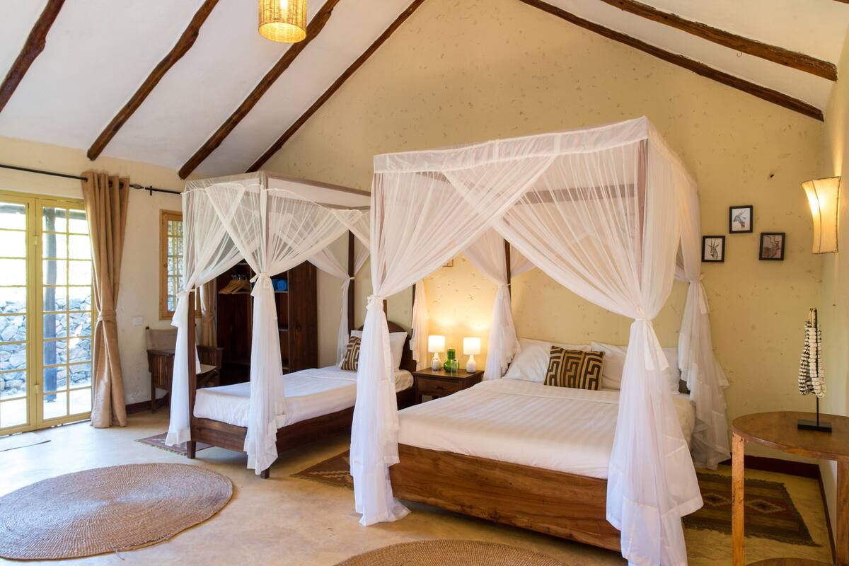 A room with two wooden beds, each with a canopy, a large window with curtains, and a round rug on the floor.