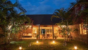 A well-lit outdoor patio area with a dining table and chairs, surrounded by lush tropical plants and a stone wall. - Kili Villa (Arusha)