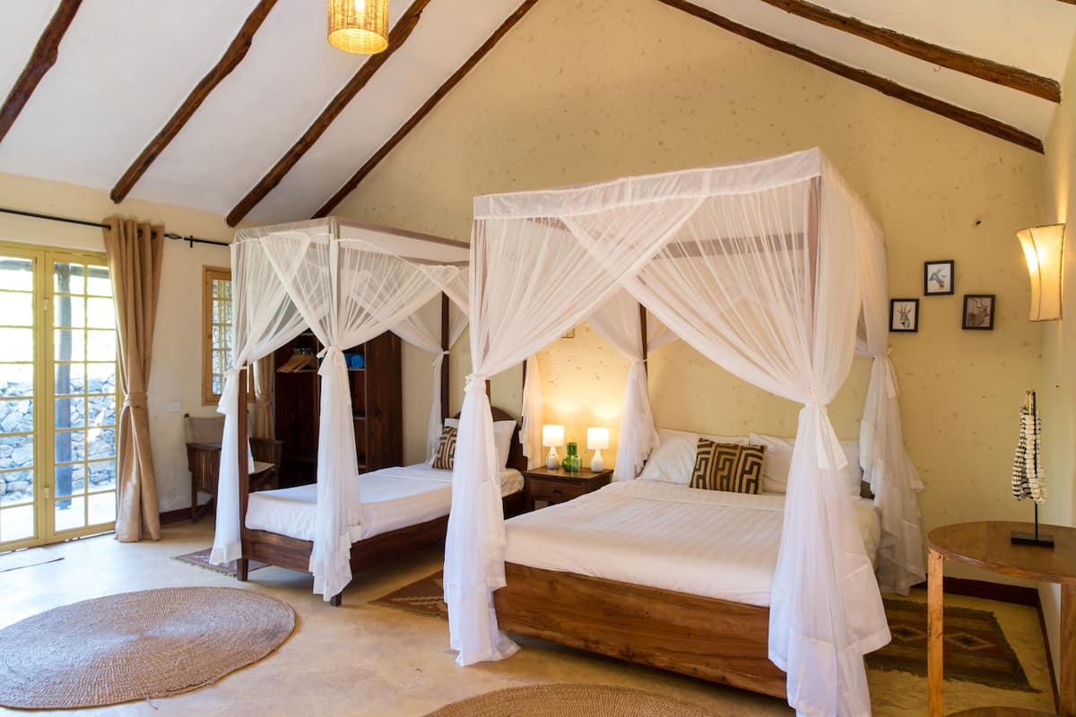 A room with two wooden beds, each with a canopy. There are two bedside tables with lamps, a large window with curtains, and a view of the outdoors.