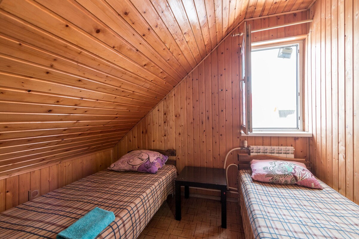 Une petite pièce chaleureuse aux murs en bois et au plafond incliné. On y trouve deux lits avec une literie à carreaux et une petite table avec des chaises. Une fenêtre laisse entrer la lumière naturelle.
