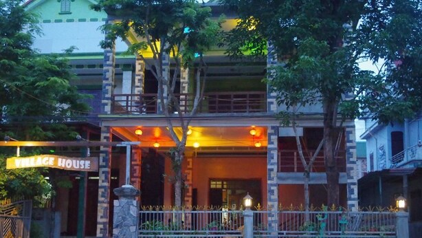 Un edificio de dos pisos con un letrero que dice "Village House". Tiene un pilar de piedra, una cerca de metal y árboles en primer plano.