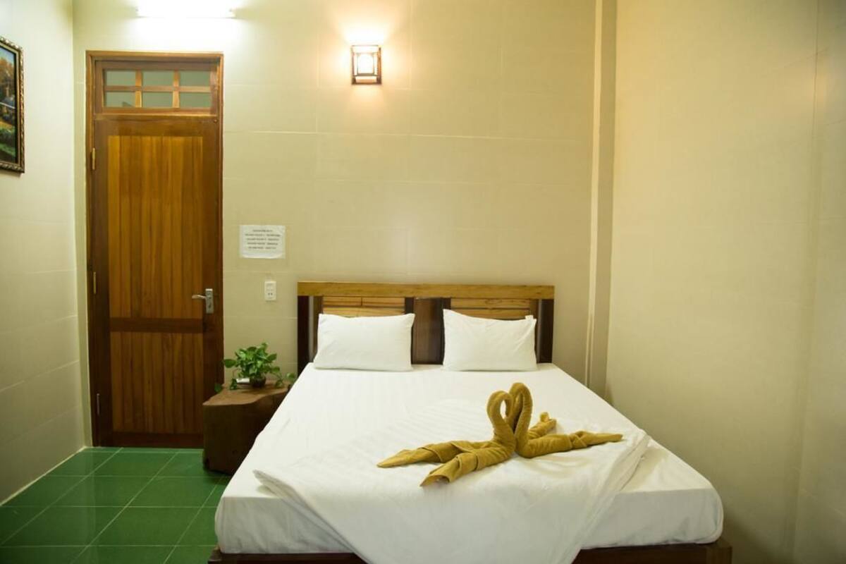 A hotel room with a wooden door, a bedside table with a plant, and a bed with a towel sculpture.