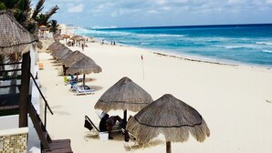 A beach with white sand, thatched umbrellas, and palm trees.
