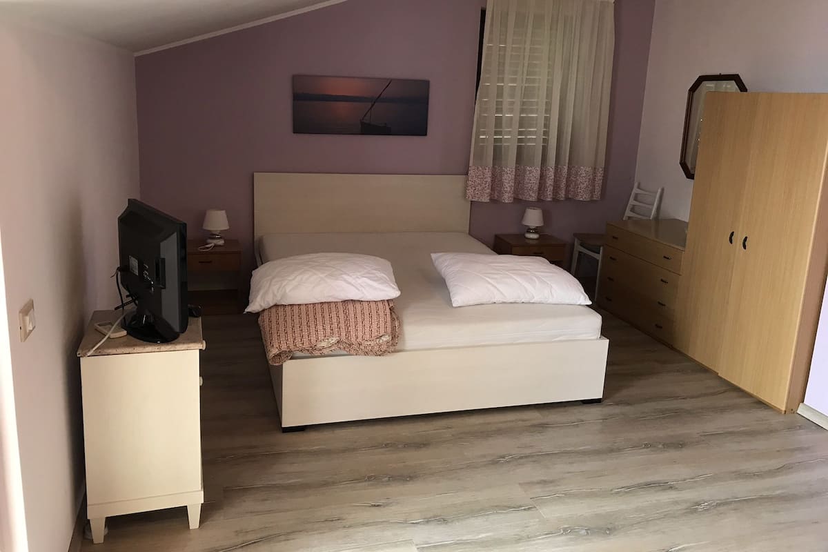 A bedroom with a bed, a television on a stand, a nightstand, a dresser, and a window with curtains.