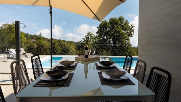 Mesa de comedor para seis personas con platos y vasos, con vista a una piscina con sillas de descanso y paisajes de árboles y colinas.