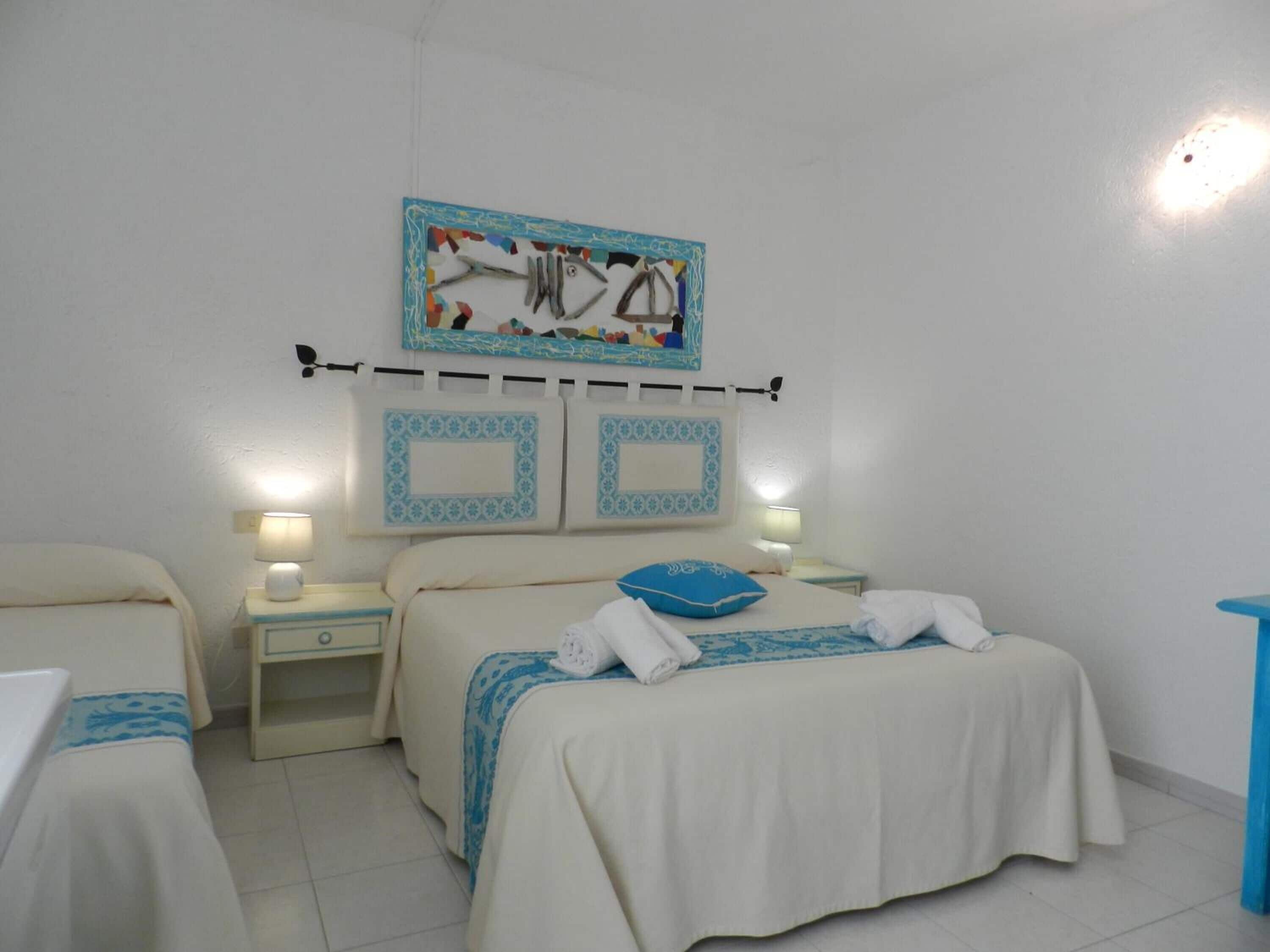Habitación de hotel con dos camas, una mesita de noche con lámpara, un cuadro en la pared y una silla azul.