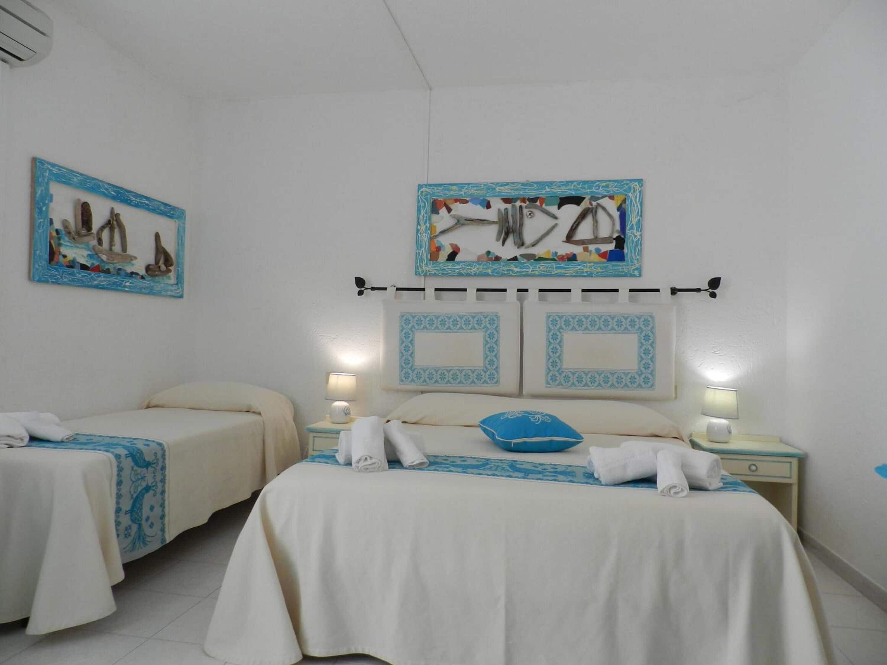 Habitación de hotel con dos camas, cada una con cubrecama blanco y almohadas decorativas azules. Hay mesitas de noche fijadas a la pared con lámparas y un adorno encima de las camas.