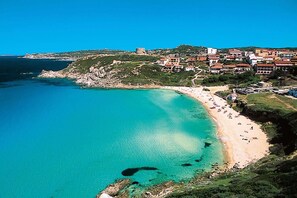 Un pueblo costero con una playa arenosa, aguas turquesas cristalinas y un castillo histórico en una colina.
