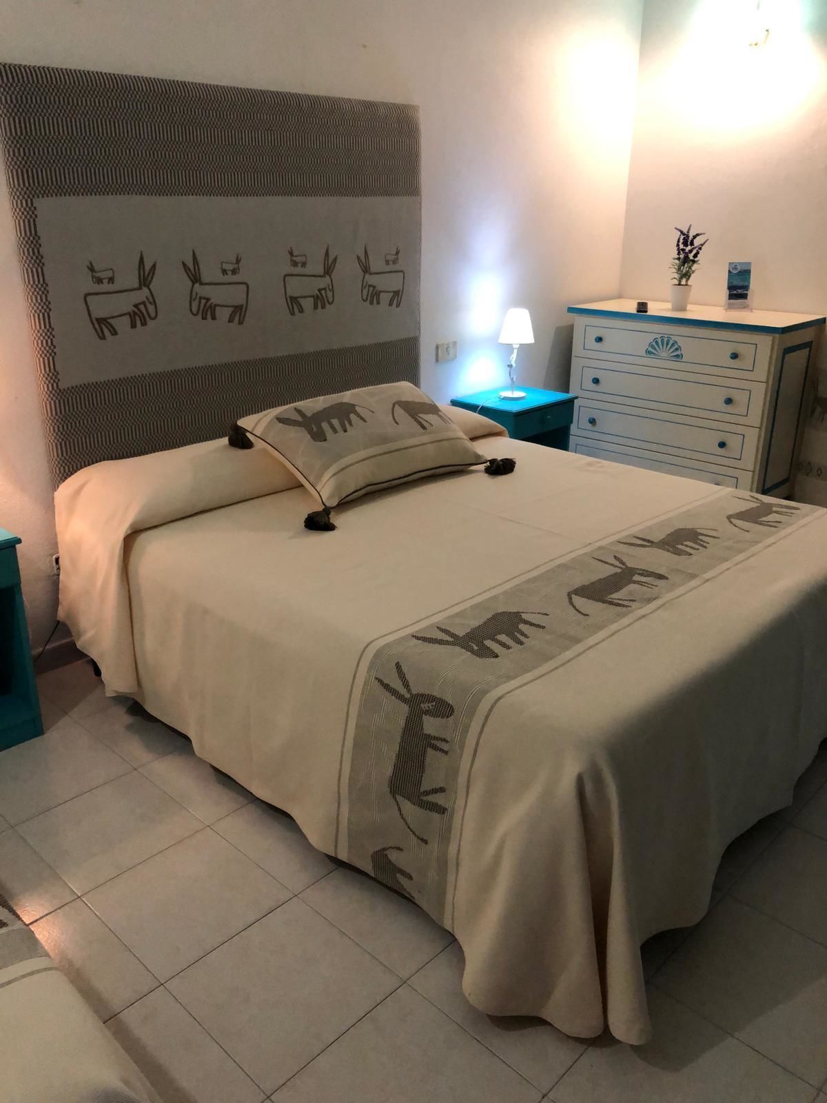 A neatly made bed with a beige blanket and pillows, a bedside table with a lamp, and a dresser with drawers.