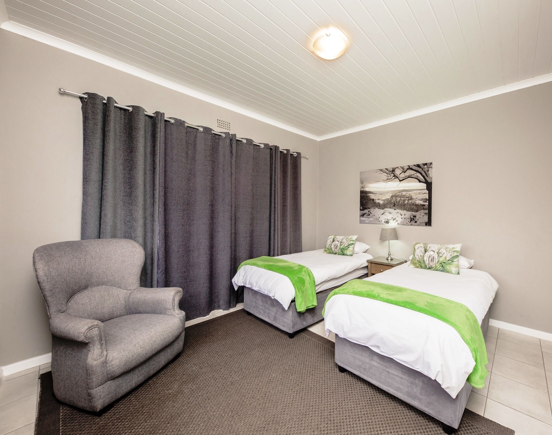 A hotel room with two beds, a grey armchair, a painting on the wall, and a ceiling light.